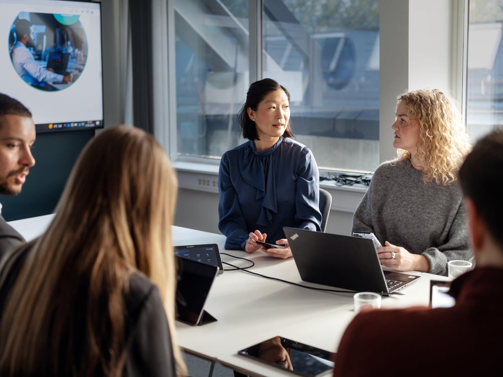 Medarbejdere engageret i en diskussion, siddende ved et bord med åbne bærbare computere, fremmer teamwork og samarbejde.
