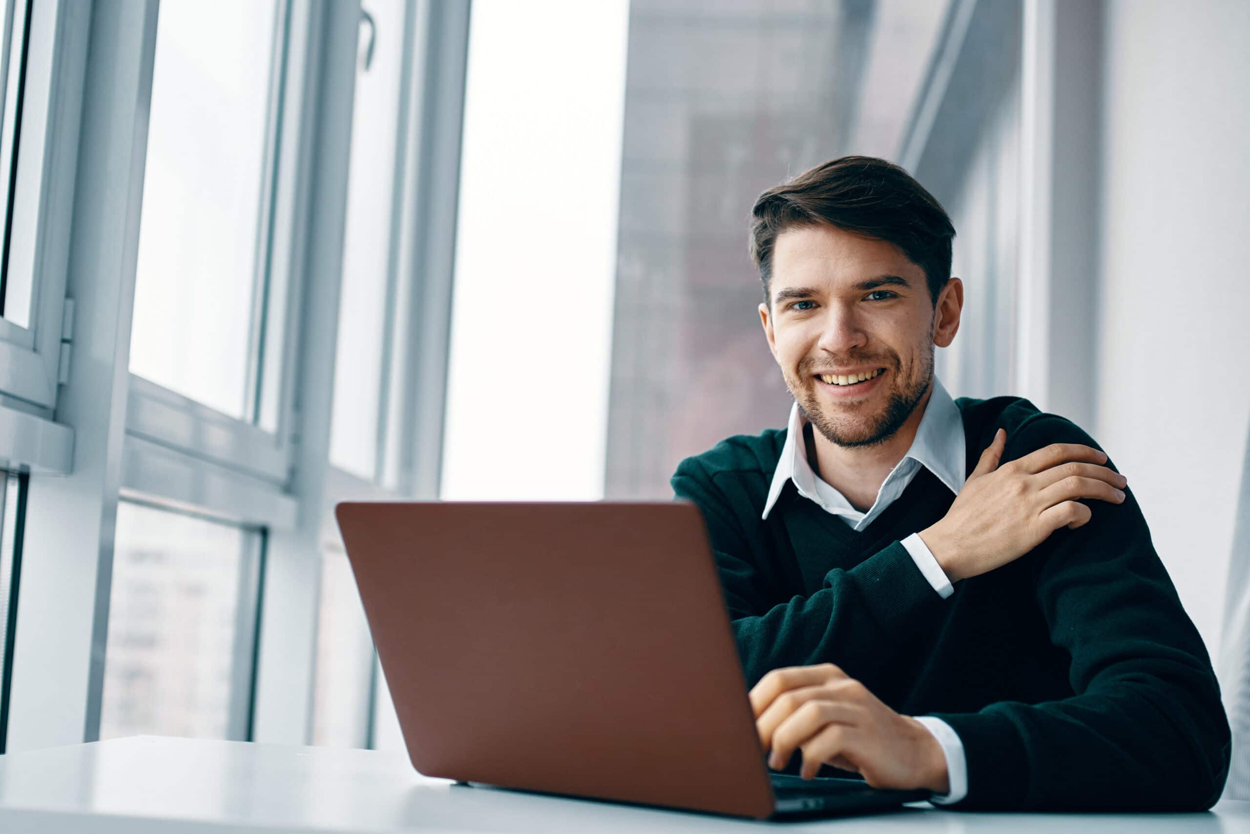 Smilende mann i mørk genser og hvit skjorte som jobber på laptop ved store kontorvindu med naturlig lys.