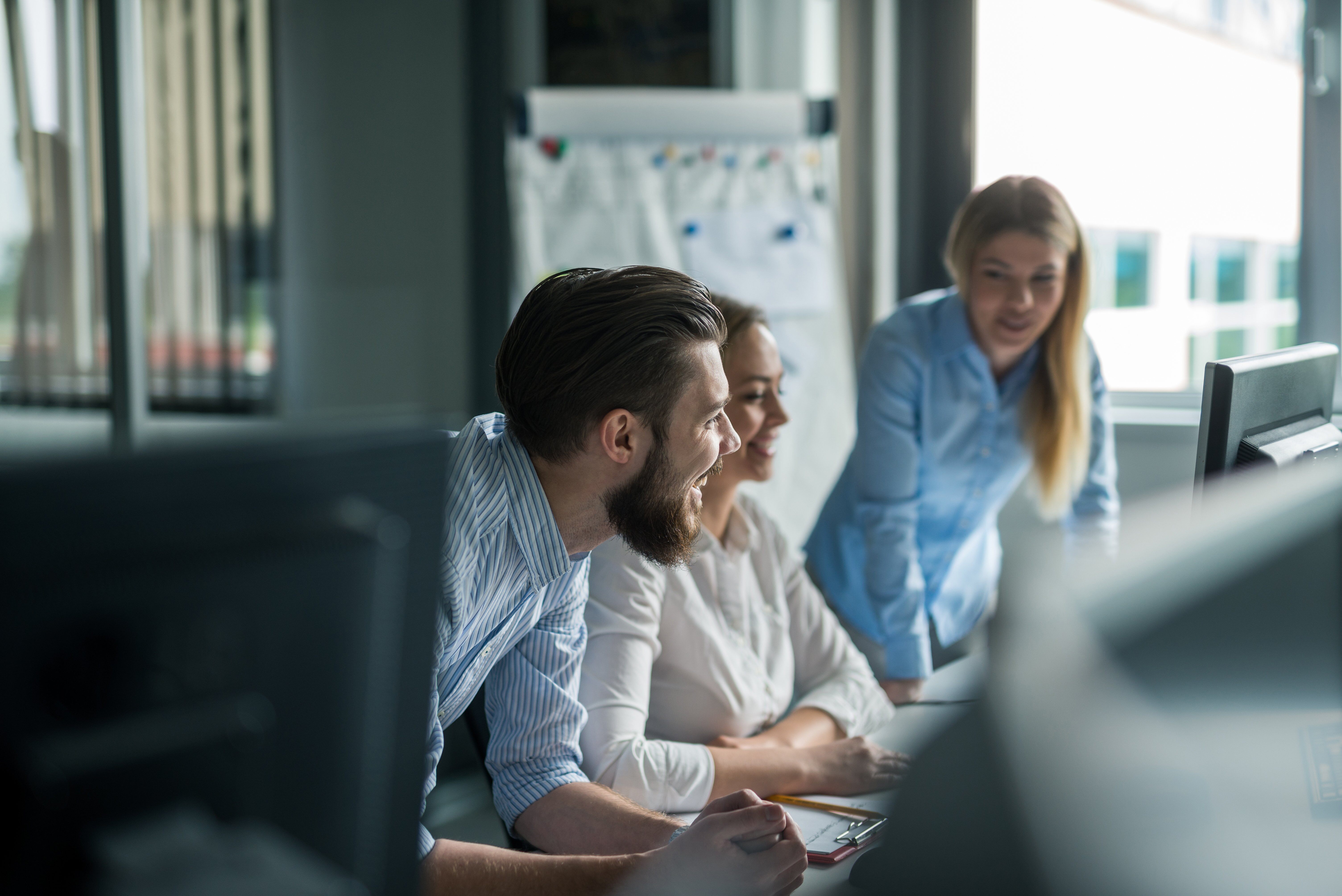 Three colleagues in an office space focused on a computer display, engaged in discussion.