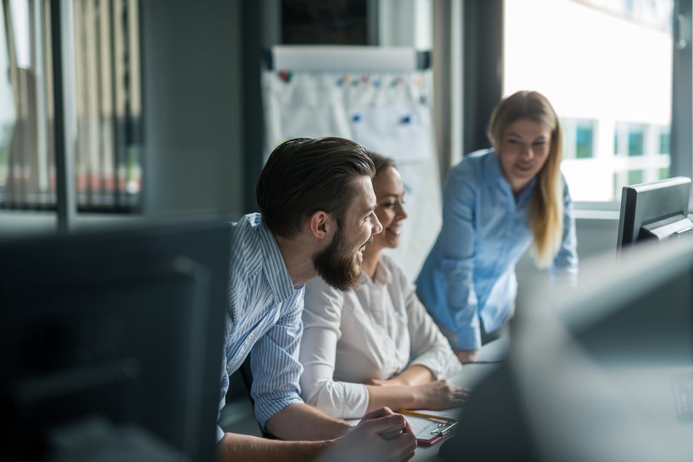 Three colleagues in an office space focused on a computer display, engaged in discussion.