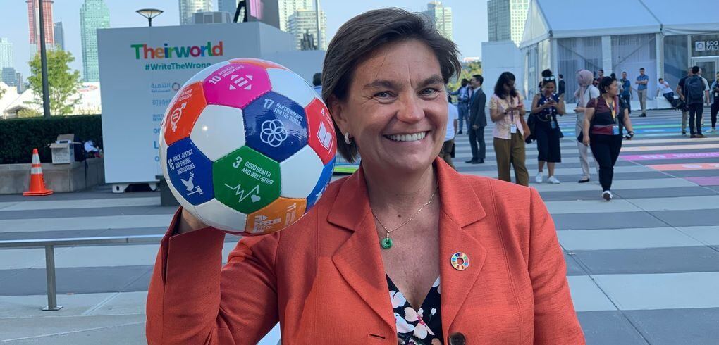 Smilende kvinne i oransje blazer holder en fotball med ikoner for FNs bærekraftsmål (SDGs)