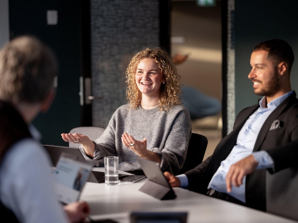 The image shows a business meeting in a modern office space where several people are seated around a table and are talking together.