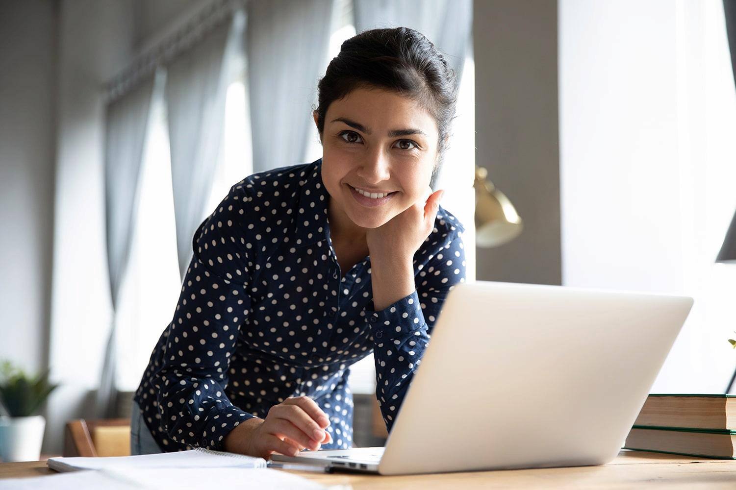 Smiling woman in a polka dot shirt leaning on a desk while working on a laptop.