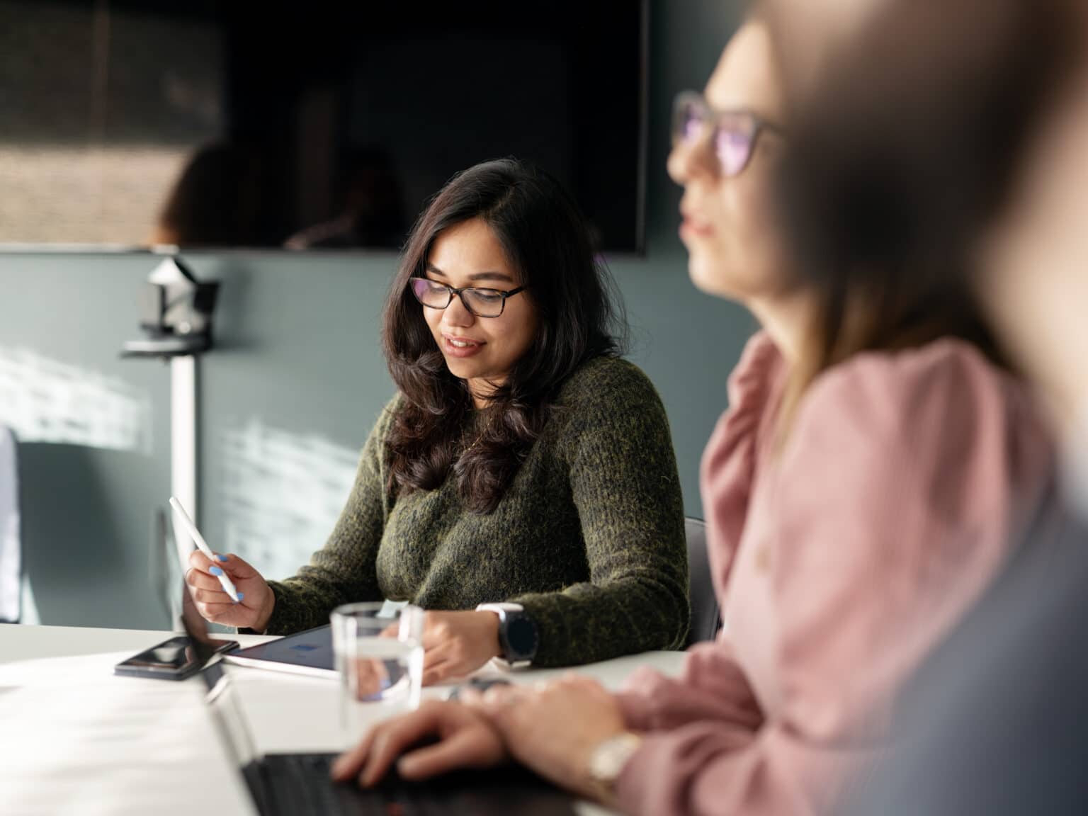 . A woman sits at a table with two others, taking notes on her tablet during a meeting.