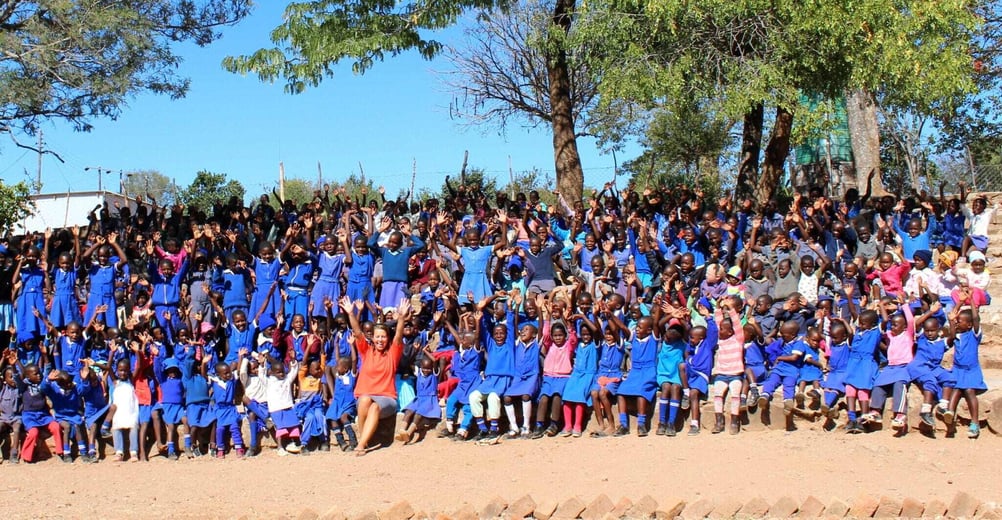 A large group of Zimbabwean school children in blue uniforms wave and smile at the camera.