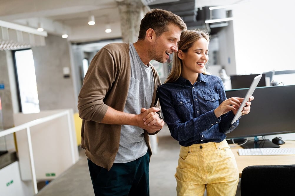 A man and a woman in an office; the woman is demonstrating something on her tablet to the man.