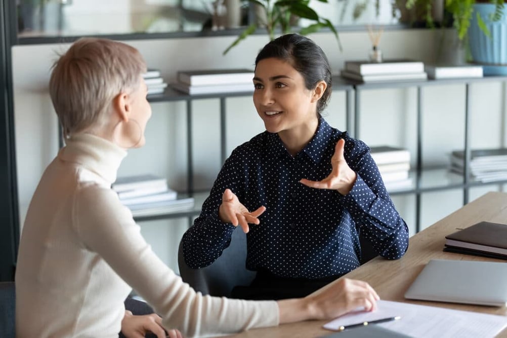 Two women engaged in conversation at a table in an office setting.