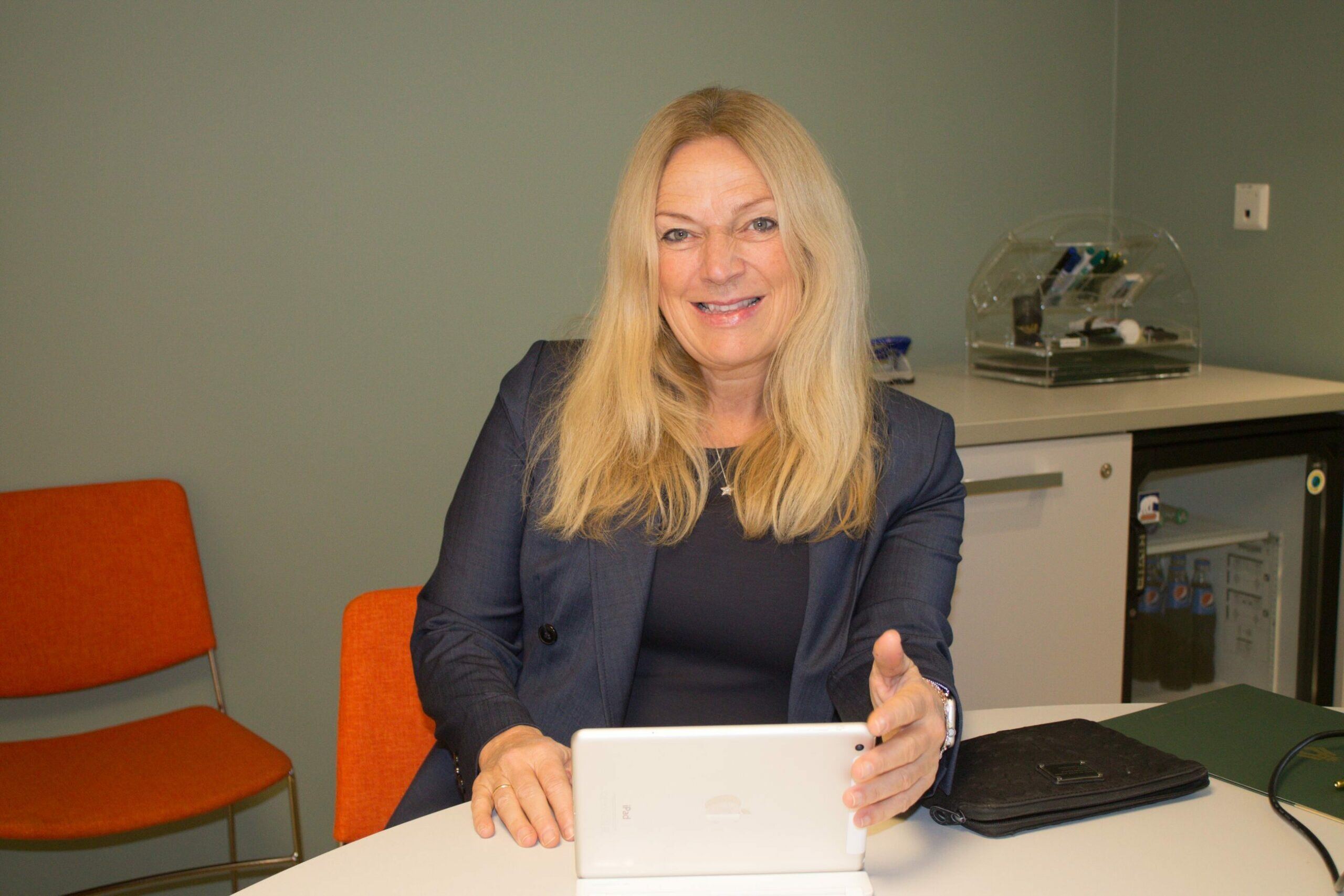 A woman in a navy blazer smiling while holding a tablet at a desk.