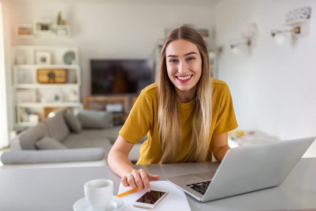 En leende kvinna sitter vid ett bord med en laptop, smartphone och kaffekopp i ett ljust, modernt vardagsrum.