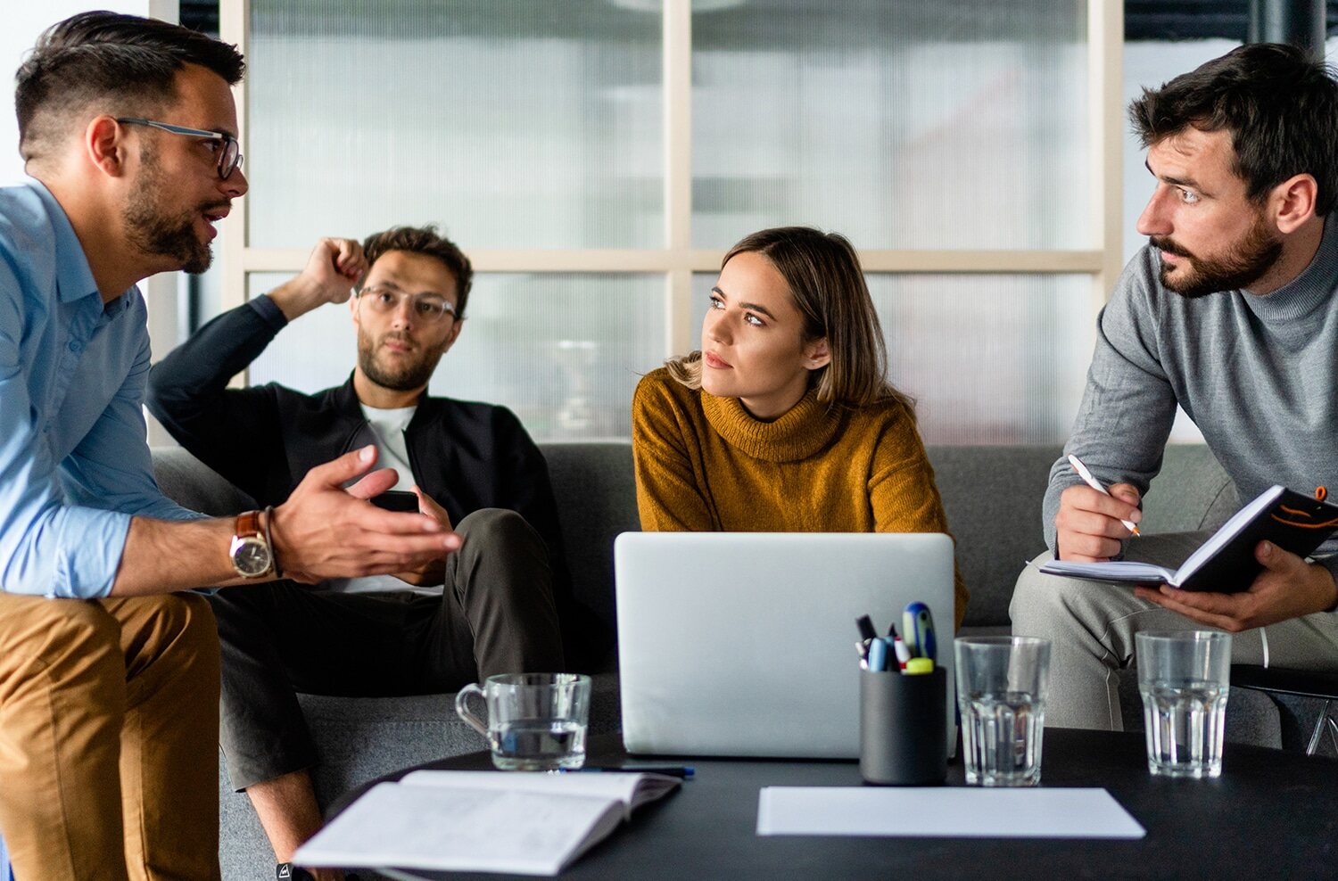 A group of four colleagues sits together in a modern office lounge and discussing. They appear engaged in a collaborative meeting