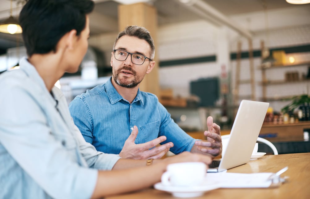 A man and woman are sitting at a table, using a laptop, appearing to collaborate or engage in a discussion.