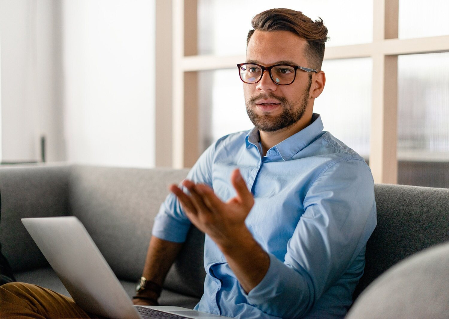 A man with glasses is seated on a couch, working on his laptop