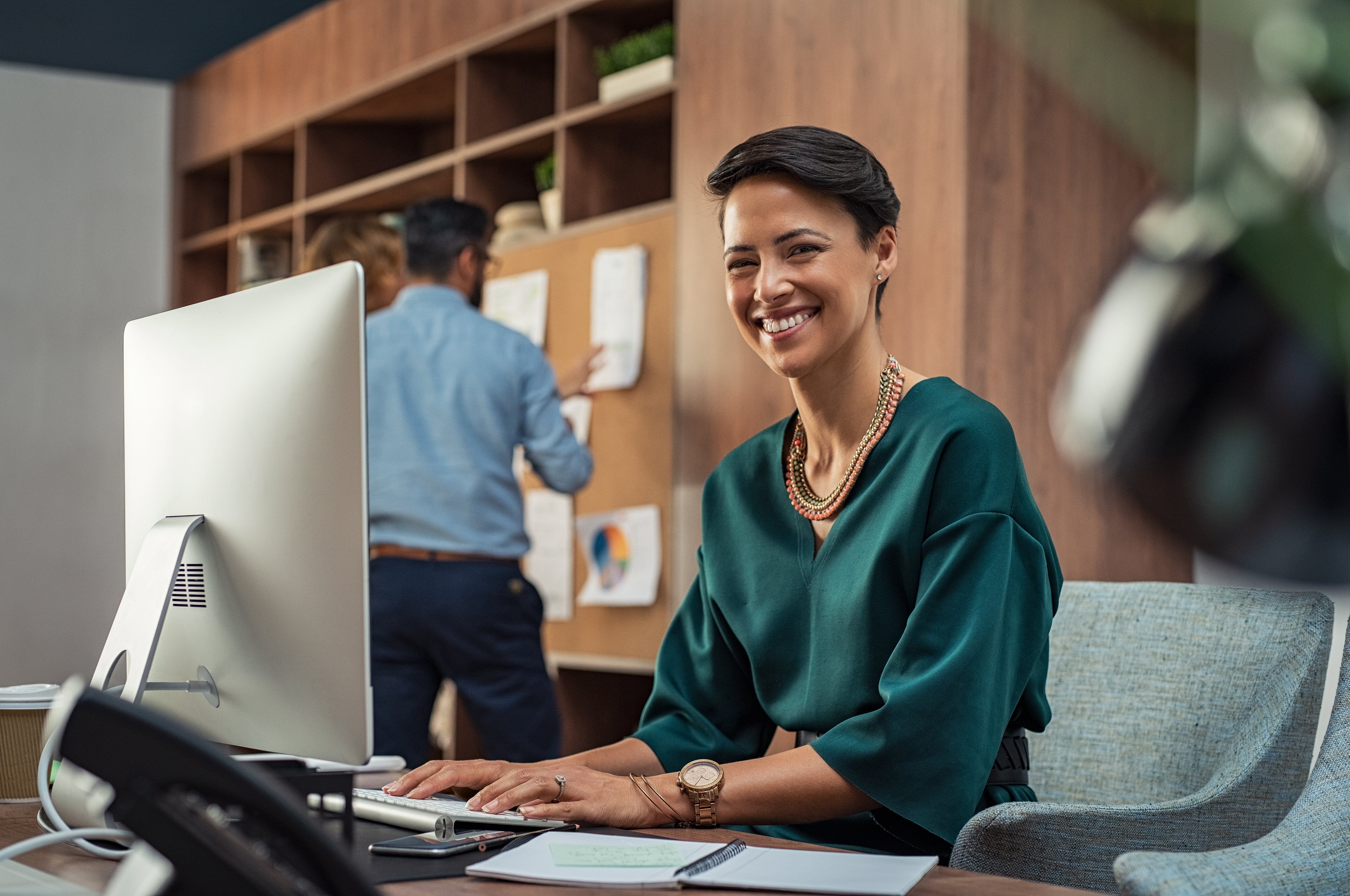 En kvinde smiler varmt, mens hun sidder ved sit skrivebord og arbejder på sin computer.