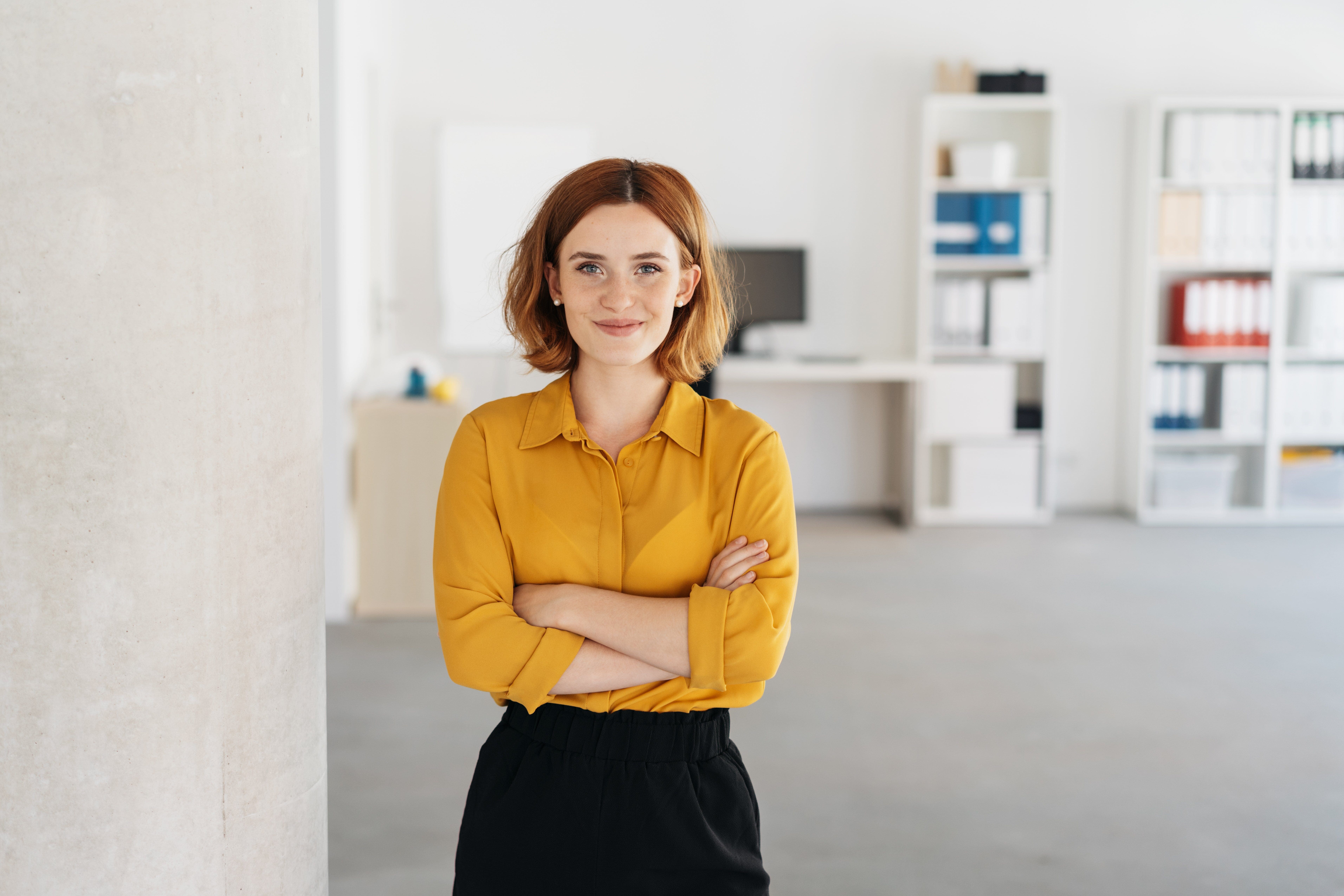 A smiling woman stands with her arms crossed in a bright office setting.