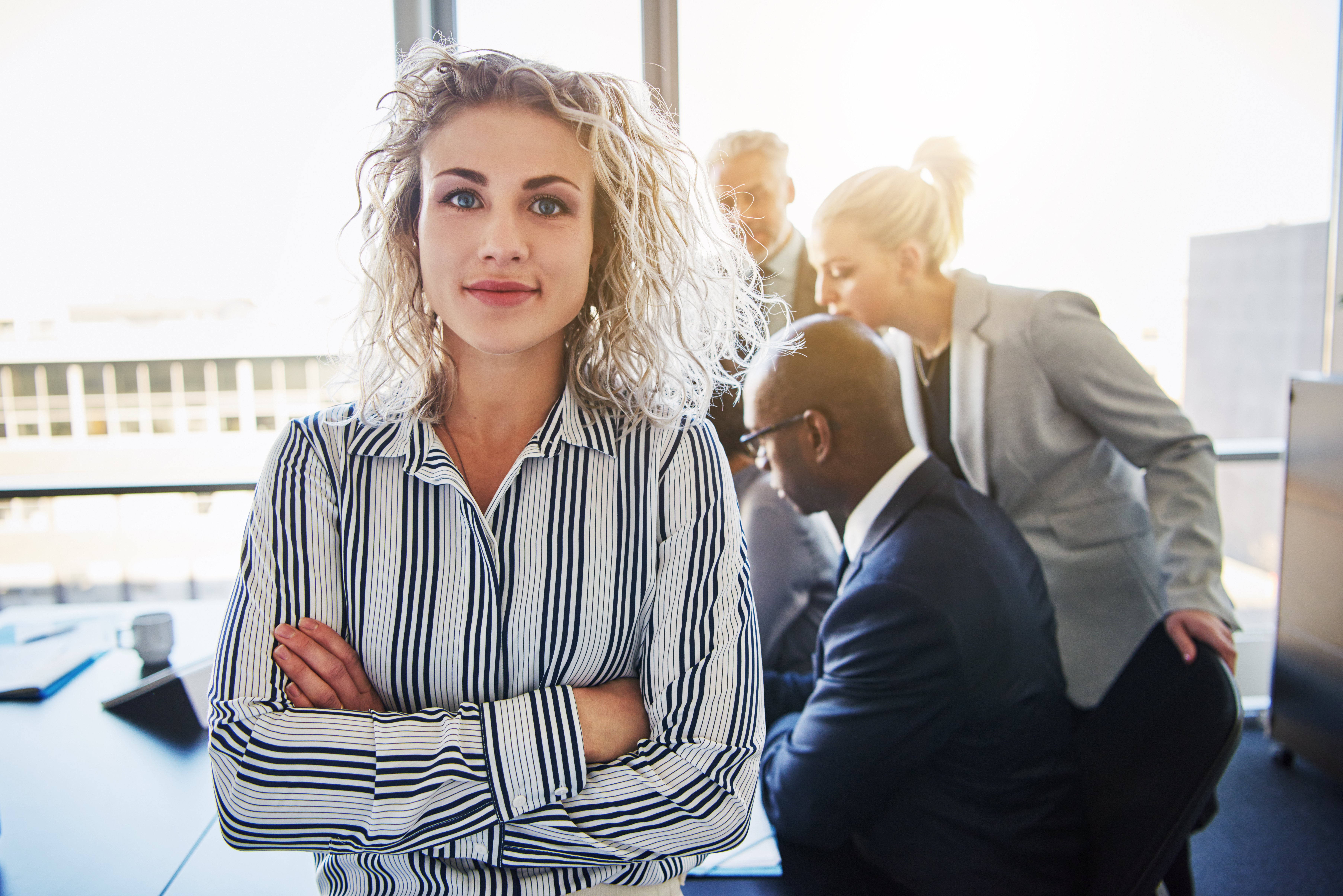 In a meeting room, a woman stands before colleagues focused on collaborating over a laptop.