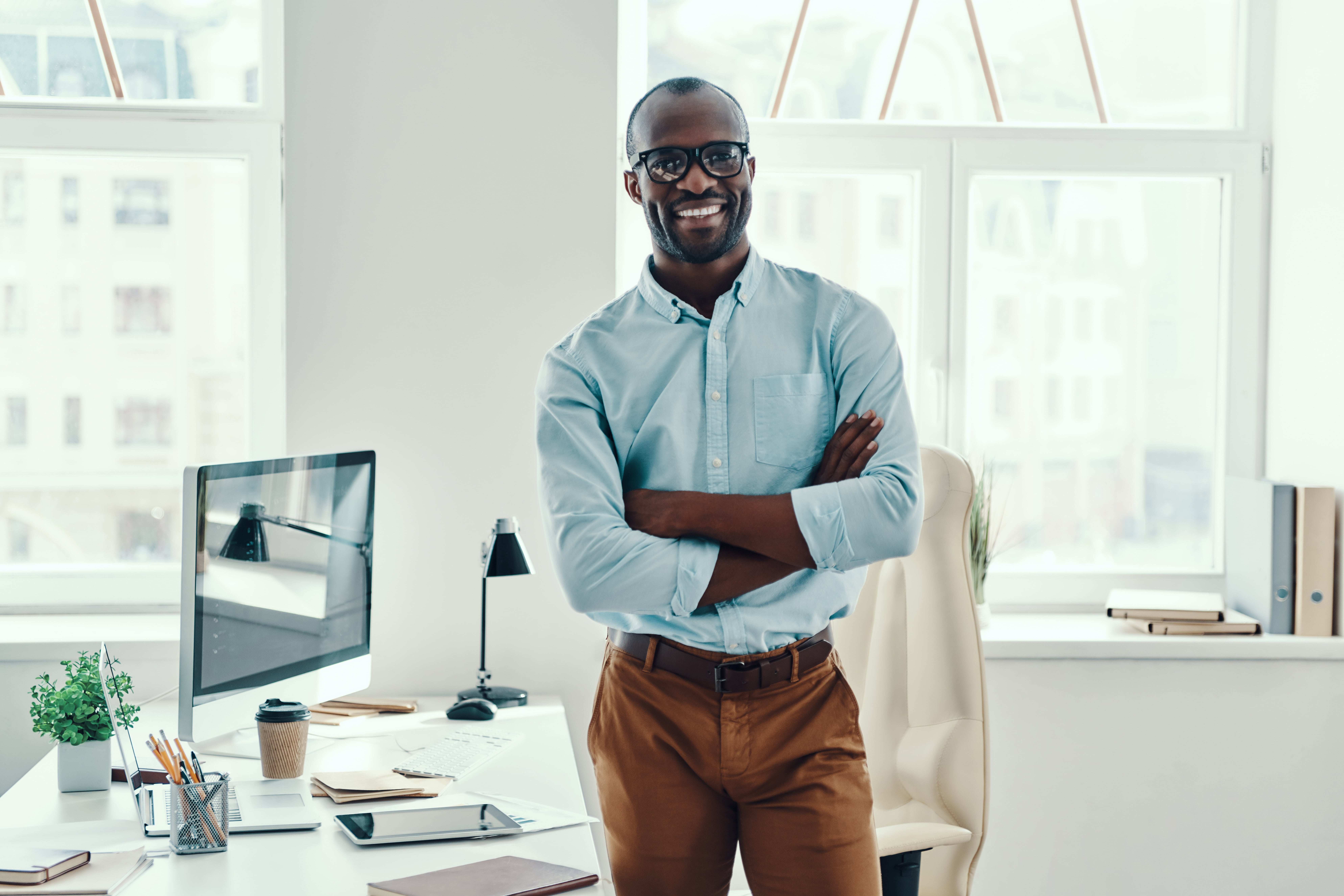 A smiling man in a light blue shirt and glasses stands with crossed arms in a bright, modern office space with a computer workstation and large windows behind him.