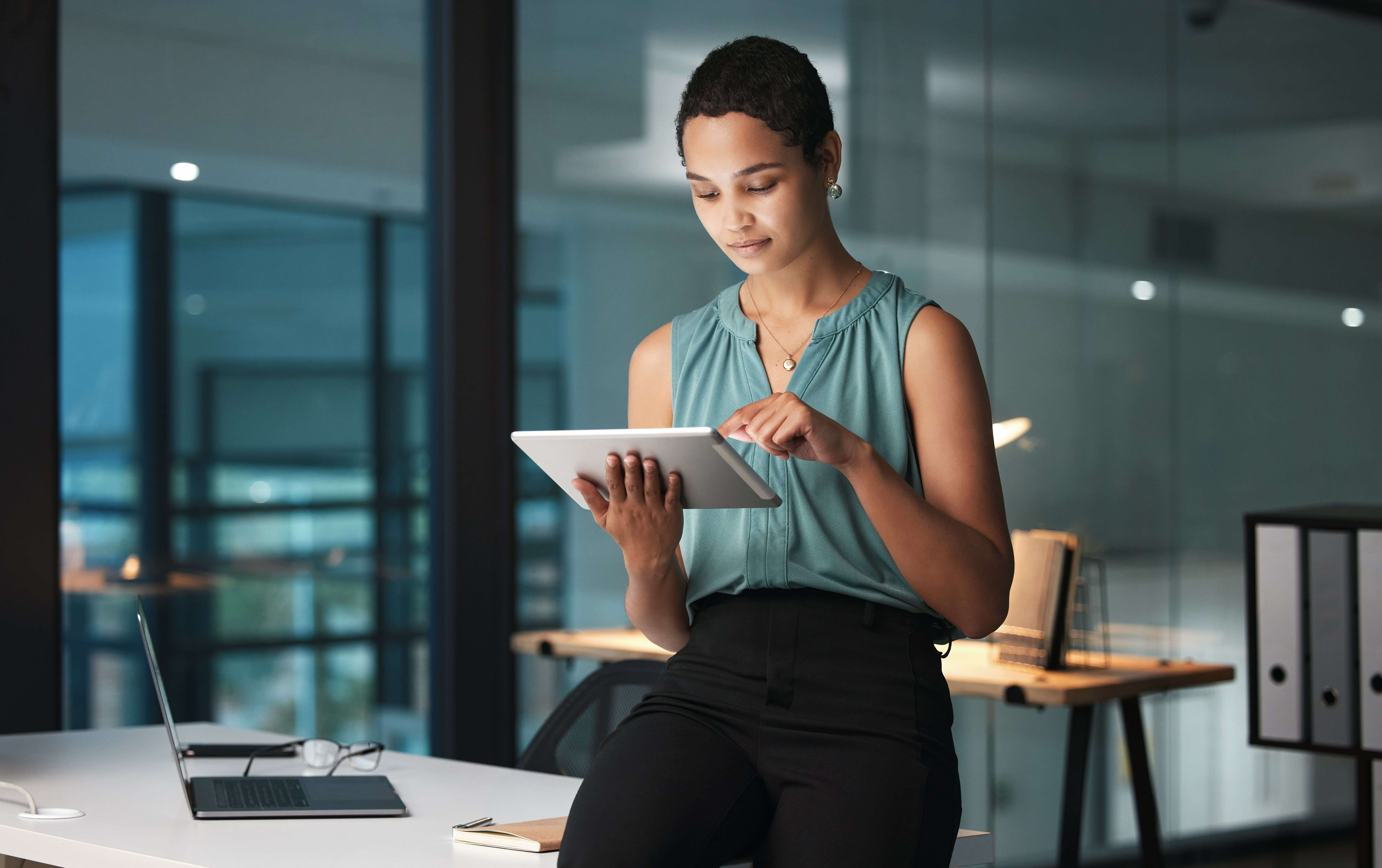 A woman reclined on a desk, engaged with a tablet, surrounded by a contemporary office environment.