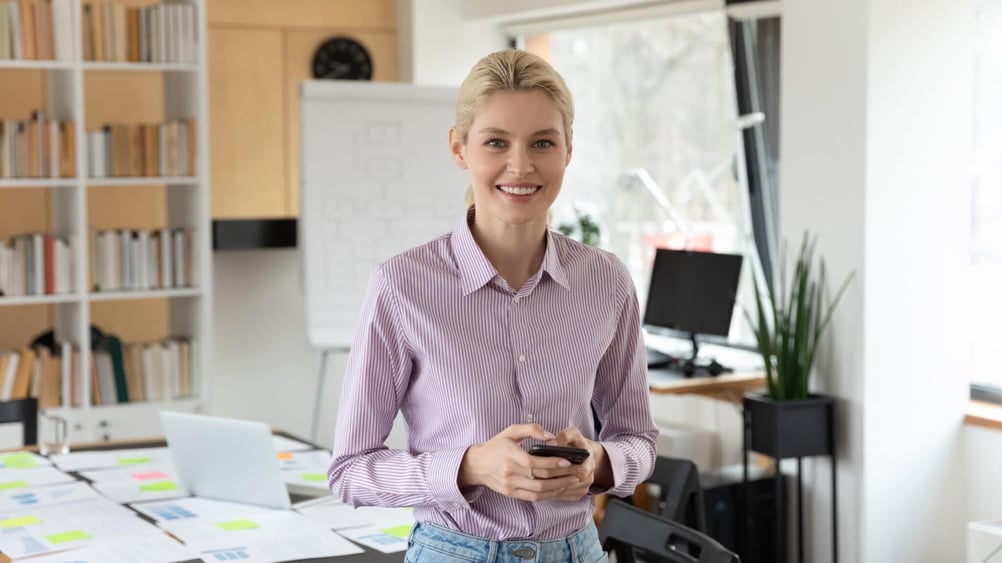 A woman stands in an office while holding a cell phone.